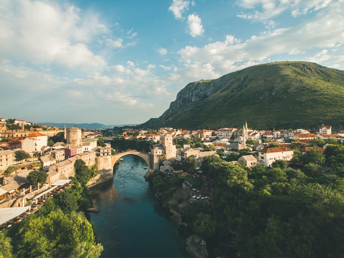 Stari Most, the stone arch bridge over the Neretva River in Mostar, with pale buildings and green trees along the riverbanks