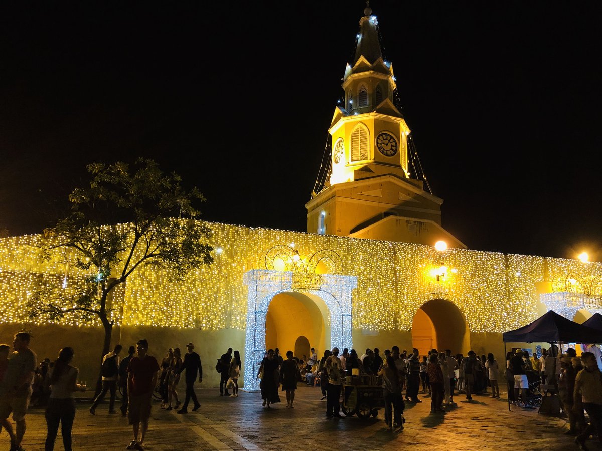 The Torre del Reloj gate and city walls of Cartagena lit up at night, with crowds in the square in front