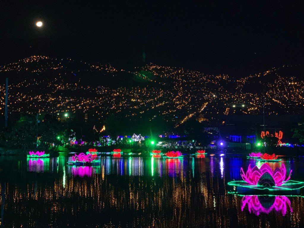 Lotus-flower light installations floating on the lake at Parque Norte, Medellín, with the hillside barrios and a full moon behind