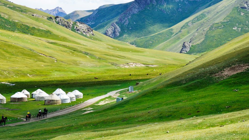 Traditional white yurts scattered across lush green alpine meadows beneath rugged mountain peaks in Kyrgyzstan, with horse riders on a dirt track through the high-altitude landscape