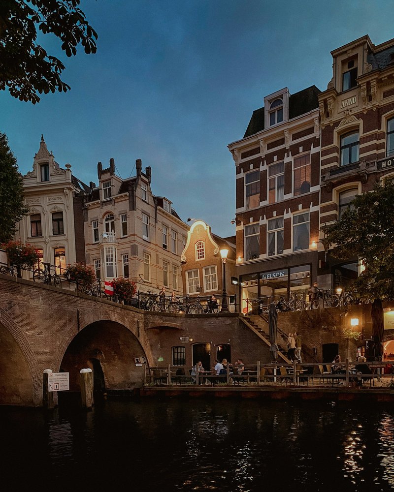 Evening view of a Utrecht canal terrace below a brick bridge, with townhouses and bicycle racks