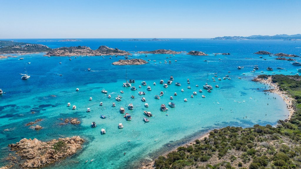 Aerial view of a turquoise bay in Sardinia, Italy, with crystal-clear Mediterranean waters, rocky islets, boats anchored offshore and a sandy beach bordered by green Mediterranean vegetation
